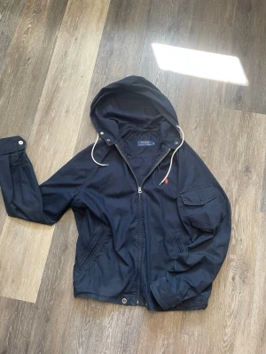 Mörkblå vindjacka med huva Polo Ralph Lauren - Snygg mörkblå vindjacka från Polo Ralph Lauren med huva och vita dragsnören. Jackan har dragkedja framtill, fickor med lock och knapp, samt en liten röd logga på bröstet. Perfekt för blåsiga dagar och enkel att matcha till olika outfits.