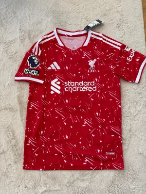 Liverpool matchtröja Szoboszlai #8 - Röd Liverpool FC matchtröja med vita detaljer och mönster, Adidas-logga och klubbmärke på bröstet. Premier League-märke på ärmen och tryck med Szoboszlai och nummer 8 på ryggen. Tillverkad i lätt och ventilerande material, perfekt för fotbollsträning eller match.