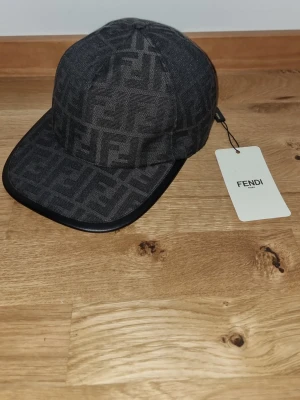 Fendi Keps - Säljer en svart keps från Fendi med det klassiska FF-logomönstret över hela kepsen. Kepsen har böjd skärm och justerbar rem baktill med Fendi Roma-detalj. Materialet är bomull och polyester, vilket gör den både snygg och bekväm.