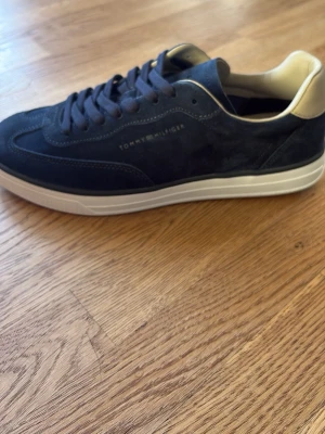 Mörkblå sneakers Tommy Hilfiger - Helt nya snygga mörkblå sneakers från Tommy Hilfiger i mocka med vit sula och beige detaljer vid hälen. Klassisk låg modell med diskret logga på sidan och matchande mörkblå skosnören. Perfekta för en stilren och avslappnad look. Dem har aldrig använts bara vid provning av storleken.