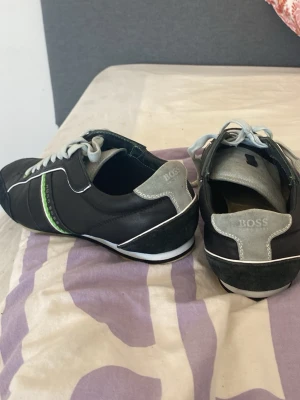 Svarta Hugo Boss sneakers med gröna detaljer - Snygga svarta sneakers från Hugo Boss med vita och gröna detaljer på sidan samt vita skosnören. Skorna har låg profil och är tillverkade i läder med mockadetaljer och en vit sula. Perfekta för dig som vill ha en stilren och sportig look.