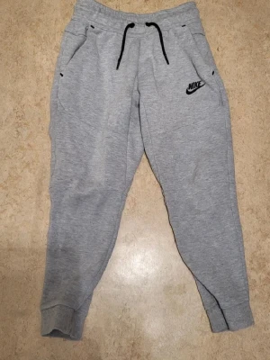 Gråa mjukisbyxor från Nike - Snygga gråa mjukisbyxor från Nike med svarta dragsnören och logga på vänster sida. Byxorna har muddar vid bensluten och två fickor fram. Perfekta för chill eller träning. Tillverkade i mjukt sweatshirtmaterial.