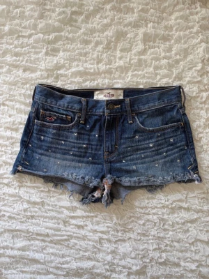 Coola Hollister jeansshorts  - Superfina mörkblå jeansshorts från Hollister i storlek W26 (motsvarar US 3). Midjemåttet rakt över är 36cm (alla mått finns i bilderna). Shortsen har snygga, slitna detaljer nertill och är dekorerade med glittrande strass-stenar på framsidan som ger en extra touch. Perfekt skick och perfekt till sommaren! ☀️