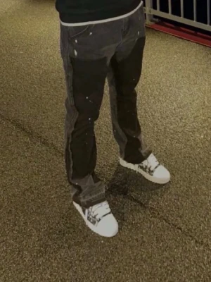 Svarta raka jeans med färgstänk - Säljer ett par svarta jeans med raka ben och råa kanter nertill. Jeansen har coola vita färgstänk över hela och en patchad detalj på ena benet. Perfekta för dig som gillar en edgy och unik stil. Materialet är klassisk denim.
