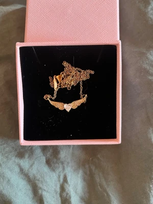 Guldpläterat halsband med vingar och hjärta - Säljer ett guldfärgat halsband med en tunn kedja och ett hänge i form av vingar med ett glittrande hjärta i mitten. Halsbandet är guldpläterat. Hänget är dekorerat med små pärlor som ger extra bling. Perfekt accessoar för dig som gillar unika detaljer och romantisk stil.