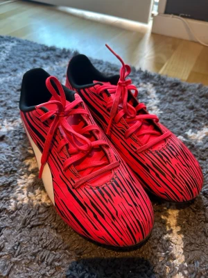 Röda Puma fotbollsskor med svart mönster - Säljer ett par snygga fotbollsskor från Puma i rött med svarta ränder och vita detaljer på sidan. Skorna har klassisk låg profil, svarta innersulor och röda skosnören. Perfekta för dig som vill sticka ut på planen!