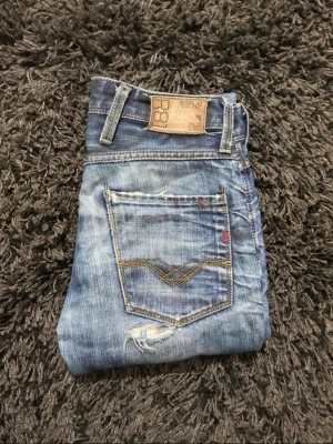Blå slitna jeans med detaljer - Säljer ett par blå jeans med slitningar och unika sömmar på bakfickan. Jeansen har en tvättad look, smal passform och coola slitdetaljer framtill. Perfekta för dig som gillar en avslappnad och trendig stil.