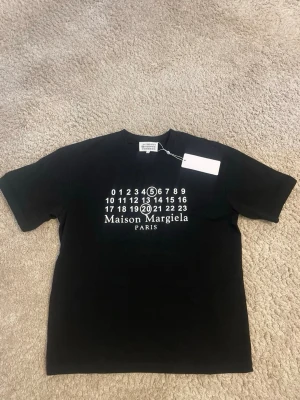 Svart Maison Margiela t-shirt - Säljer en svart t-shirt från Maison Margiela med det ikoniska numermotivet tryckt på bröstet. T-shirten har rund hals och korta ärmar. Tillverkad i mjuk bomull för en skön känsla och clean look.