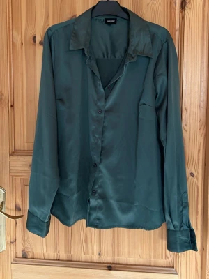 Mörkgrön satinblus med knappar - Snygg mörkgrön skjorta i satinliknande material med klassisk krage och knäppning framtill. Långärmad modell med breda manschetter och en avslappnad passform. Perfekt för dig som vill ha en stilren och trendig look.