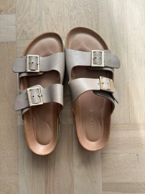 Beiga sandaler med spännen från Sweeks  - Snygga beige sandaler från Sweeks med två breda remmar och justerbara metallspännen. Fotbädden är formad för extra komfort och sulan är vit med tydlig logga undertill. Perfekta för en avslappnad och trendig look.