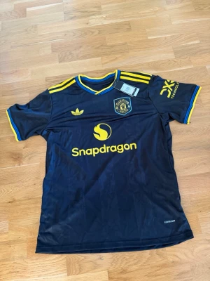 Manchester United x Adidas matchtröja - Snygg svart Manchester United matchtröja från Adidas med gula och blå detaljer. Tröjan har klubbmärke och Adidas-logga på bröstet samt Snapdragon som sponsortryck. Tillverkad i lätt och ventilerande material, perfekt för fotboll eller träning.