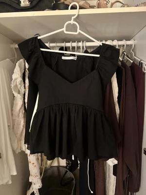 Svart topp med volang och v-ringning - Säljer en svart topp i storlek XS med v-ringad hals och volang nedtill. Toppen har korta puffärmar och är tillverkad i ett lätt, ribbat material. Perfekt för dig som gillar en feminin och trendig stil.
