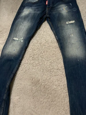 Mörkblå slitna jeans med tryck - Snygga mörkblå jeans med slitningar och ljusa partier på benen. Jeansen har smal passform, detaljer med färgstänk och ett stort rött 'ICON2'-tryck på baksidan. Perfekta för en trendig streetstyle-look.