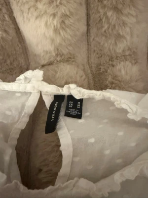 Vit blus med volanger från Vero Moda - Söt vit blus från Vero Moda i transparent, prickigt tyg. Blusen har volangdetaljer vid ärmarna och spets längs bröstet. Luftig och lätt med rund halsringning och korta ärmar. Perfekt för en trendig och romantisk look.
