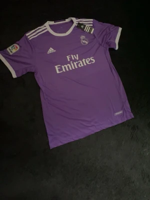 Lila Real Madrid Ronaldo matchtröja - Säljer en lila Real Madrid matchtröja från Adidas med vita detaljer och tryck. Tröjan har Ronaldo och nummer 7 på ryggen, samt Fly Emirates-logga på bröstet. Korta ärmar och klassisk sportig passform. Perfekt för fotbollsfans!