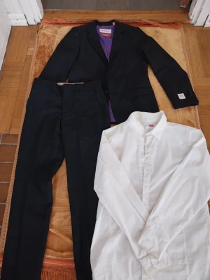 Svart kostym från Opposuits - Snygg svart kostym från Opposuits med lila innerfoder och klassisk tvåknäppt kavaj. Tillhörande byxor i samma färg och en vit skjorta med knappar. Perfekt för dig som vill ha en stilren och modern look. Stl146-152.använd 2gånger