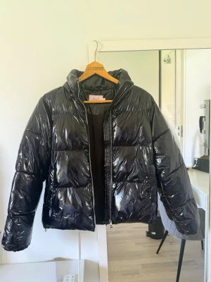 Svart glansig pufferjacka från Nelly - Snygg svart pufferjacka från Nelly som är glansig. Jackan har dragkedja framtill, huva och breda ärmar. Perfekt för kalla vinterdagar och ger en trendig look till din outfit. Jackan är aldrig använd och därför i topp skick.