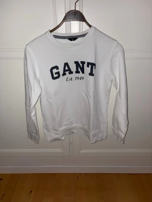 Vit sweatshirt GANT - Klassisk vit sweatshirt från GANT med rund halsringning och tryckt logga i mörkblått framtill. Tröjan har långa ärmar och ribbade muddar vid ärmslut och nederkant. Perfekt för en avslappnad och stilren look. 