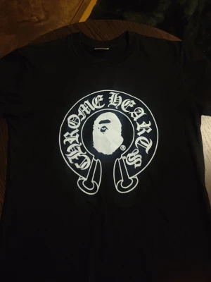 Svart Chrome Hearts x BAPE T-shirt - Säljer en svart t-shirt med stor vit Chrome Hearts och BAPE Baby Milo-print både fram och bak. T-shirten har klassisk passform och korta ärmar. Materialet känns som mjuk bomull och trycket är riktigt tydligt och snyggt.