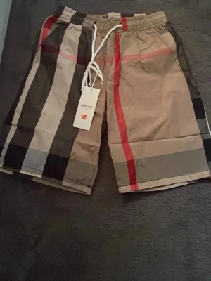 Beige rutiga shorts med snörning - Säljer ett par beige shorts med stora rutor i svart, vitt, grått och röda detaljer. Shortsen har snörning i midjan och elastisk linning för skön passform. Perfekta för sommaren och har en modern, street-inspirerad look.