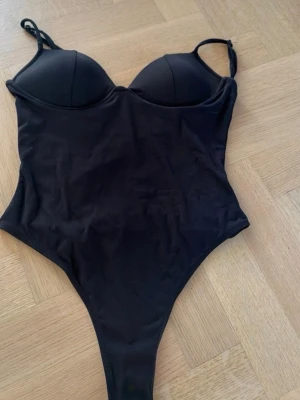 Populär body - Stilren svart body med vadderade kupor och justerbara axelband. Tillverkad i ett stretchigt, mjukt material som formar sig efter kroppen. Kuporna har en ljus insida och ger bra stö. Perfekt under tighta plagg för en slät siluett.