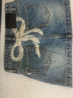 Kort jeanskjol med bakfickor - Säljer en blå kort jeanskjol med två bakfickor och en vit sprayad detalj på baksidan. Perfekt för dig som gillar en avslappnad och trendig stil.