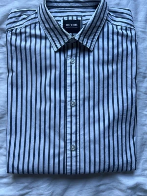 Randig skjorta från Only & Sons - Säljer en vit skjorta med mörkblå vertikala ränder från Only & Sons. Skjortan har klassisk krage, knappar framtill och regular fit. Perfekt för dig som gillar stilrena och tidlösa plagg.