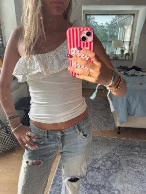 Vit oneshoulder topp med volang - Säljer en vit oneshoulder topp med bred volang i broderad spets. Toppen är tight och ärmlös, perfekt för dig som vill ha en unik och somrig look. Passar till både jeans och kjol för en trendig outfit.