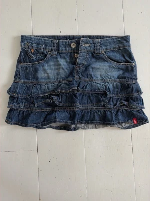 Jeanskjol med volanger från Levi's - Säljer en blå jeanskjol från EDC med flera volanglager och klassiska jeansdetaljer som fickor fram och bak samt bälteshällor. Kjolen har en kort passform och stängs med knapp och dragkedja framtill. Perfekt för en trendig och avslappnad look.