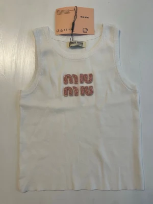 Vit ribbad linnetopp från Miu Miu - Säljer en vit ribbad linnetopp från Miu Miu med rosa glittrig text framtill. Toppen är ärmlös och har en klassisk rund halsringning. Perfekt för dig som gillar en enkel men ändå trendig look.