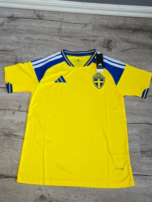 Svensk landslagströja Adidas 2026 - Gul och blå fotbollströja från Adidas med Sveriges landslagsemblem på bröstet. Tröjan har korta ärmar, blå detaljer på axlar och ärmslut samt 'Sverige' tryckt i nacken. Tillverkad i lätt och ventilerande material.