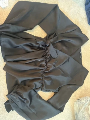 Svart omlottblus från Missay - Snygg svart omlottblus från Missay i glansigt material. Blusen har långa puffärmar med resår vid ärmslut och knytdetalj i midjan. Perfekt för dig som gillar trendiga och stilrena plagg med en twist.