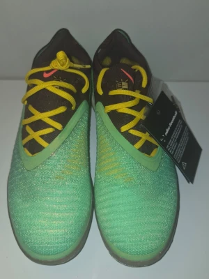 Nike gröna fotbollsskor med gula snören - Snygga fotbollsskor från Nike i mintgrönt med gula snören och bruna detaljer. Skorna har en låg profil och är tillverkade i ett stickat material för bra passform och känsla på planen. Swoosh-logga på sidan och tag på plösen. Perfekta för inomhusspel.