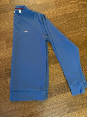 Blå sweatshirt från Gant - Snygg blå sweatshirt från Gant med broderad logga på bröstet. Tröjan har rund halsringning, långa ärmar och ribbade muddar vid ärmslut och nederkant. Perfekt för en avslappnad och stilren look.
