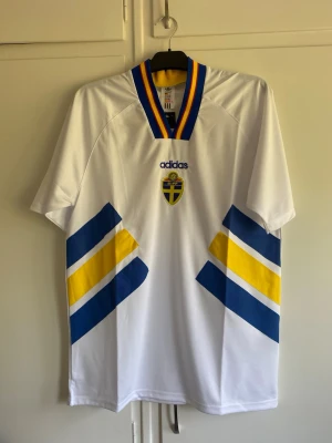 Svensk landslagströja Adidas vit - Säljer en vit svensk landslagströja från Adidas med blå och gula detaljer på framsidan. Tröjan har korta ärmar, V-ringad krage med blå och gula ränder samt Sveriges landslagsemblem på bröstet. Perfekt för fotbollsfans!