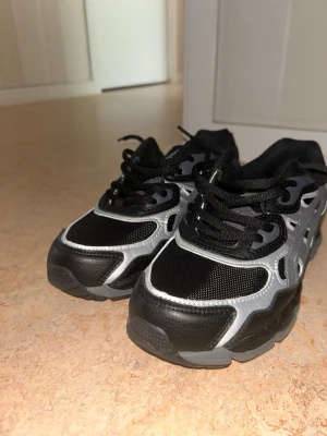 Svarta och grå Asics Gel - Aldrig använda Snygga sneakers från Asics i svart och grått med coola detaljer. Skorna har meshpaneler för bra ventilation och partier i syntetläder. Klassisk snörning och låg profil gör dem perfekta för en sportig look.