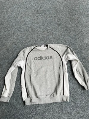 Grå och vit Adidas sweatshirt - Snygg grå sweatshirt från Adidas med vita paneler längs ärmarna och svarta detaljer. Stor Adidas-logga framtill. Tröjan har rund halsringning, långa ärmar och ribbade muddar. Perfekt för en sportig och avslappnad stil.