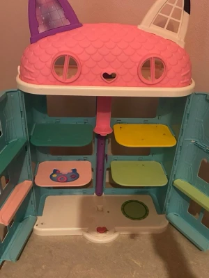 Färgglatt dockhus med kattöron - Säljer ett lekfullt dockhus i plast med kattöron och fönster, i färgerna rosa, vitt, blått, grönt och gult. Huset har flera våningar och öppningsbara dörrar samt en tillhörande lekplats med gunga och rutschkana. Perfekt för fantasifull lek! Köpt för 3000kr säljer för 1000kr