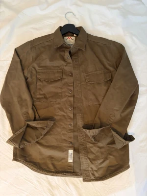 Olivgrön overshirt från Vimanron - Stilren olivgrön overshirt från Vimanron med klassisk krage och två bröstfickor med lock. Skjortan har knäppning framtill och är tillverkad i ett slitstarkt bomullstyg. Perfekt att bära öppen eller stängd för en avslappnad look.