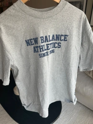 Grå New Balance Athletics t-shirt - Snygg grå t-shirt från New Balance med blå text 'NEW BALANCE ATHLETICS SINCE 1906' på bröstet. Klassisk rund halsringning och korta ärmar. Tillverkad i mjukt bomullsmaterial som känns skönt mot huden.