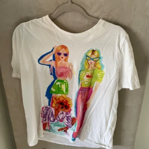 Vit t-shirt med färgglatt tryck - Vit t-shirt med coolt och färgstarkt tryck av tre tecknade personer på framsidan. T-shirten har rund hals och korta ärmar, perfekt för dig som gillar unika och lekfulla plagg. Materialet känns mjukt och skönt mot huden.