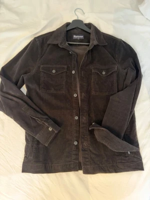 Brun manchester overshirt från Barbour - Snygg brun overshirt från Barbour i mjuk manchester. Klassisk skjortkrage, två bröstfickor med knapp och knäppning framtill. Perfekt lager-på-lager-plagg med avslappnad passform och stilrena detaljer.