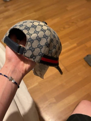 Beige keps med GG-mönster  - Snygg beige keps från med klassiskt GG-monogram över hela kepsen och en röd-blå rand på sidan. Justerbar rem baktill i mörkblått läder och böjd skärm. Perfekt accessoar för en trendig och exklusiv look.