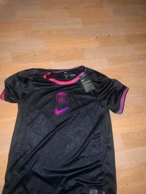 Svart PSG x Nike fotbollströja - Snygg svart PSG fotbollströja från Nike med rosa detaljer på krage och ärmslut. Tröjan har PSG-logga och Nike Swoosh i rosa framtill samt diskret blommönster i tyget. Perfekt för träning eller match.