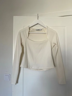 Vit långärmad topp från NA-KD - Säljer en stilren vit långärmad topp från NA-KD. Toppen har en croppad passform och en unik skärning vid halsen som ger en modern look. Perfekt att matcha med både jeans och kjol för en trendig outfit.