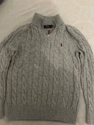 Grå kabelstickad tröja Polo Ralph Lauren - Kabelstickad grå tröja från Polo Ralph Lauren med halv dragkedja och hög krage. Tröjan har ribbade muddar och nederkant samt den klassiska broderade loggan på bröstet. Perfekt för lager-på-lager och chill stil.