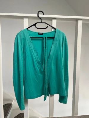 Gerry Weber kofta - Snygg turkos omlottkofta från Gerry Weber Edition med flätat knytband i midjan. Koftan har djup V-ringning och långa ärmar. Perfekt att slänga över en topp för en fräsch look. Materialet känns mjukt och följsamt. Nypris ca 1300. Storlek 34 så passar S/XS