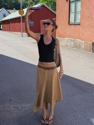 Beige långkjol med bälte och asymmetrisk fåll - Snygg beige långkjol med asymmetrisk fåll och dekorativt bälte i midjan. Kjolen har en lätt A-linjeform och är perfekt för dig som gillar en avslappnad men trendig stil. Materialet ser ut att vara linne eller bomull, vilket gör den luftig och skön.