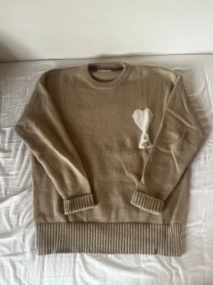 Beige stickad Ami Paris tröja  - Beige stickad tröja med rund halsringning och ribbade muddar. På bröstet finns en vit hjärtdetalj med bokstaven A under. Tröjan har en avslappnad passform och är perfekt för lager på lager under kyliga dagar. Säljer eftersom att den inte används. Storlek M 