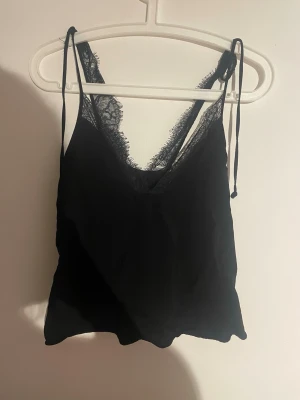 Svart linnetopp med spetsdetaljer - Snygg svart linnetopp med tunna axelband och v-ringning. Toppen har fina spetsdetaljer längs urringningen och är tillverkad i ett lätt och mjukt material. Perfekt att styla med jeans eller kjol för en trendig look.